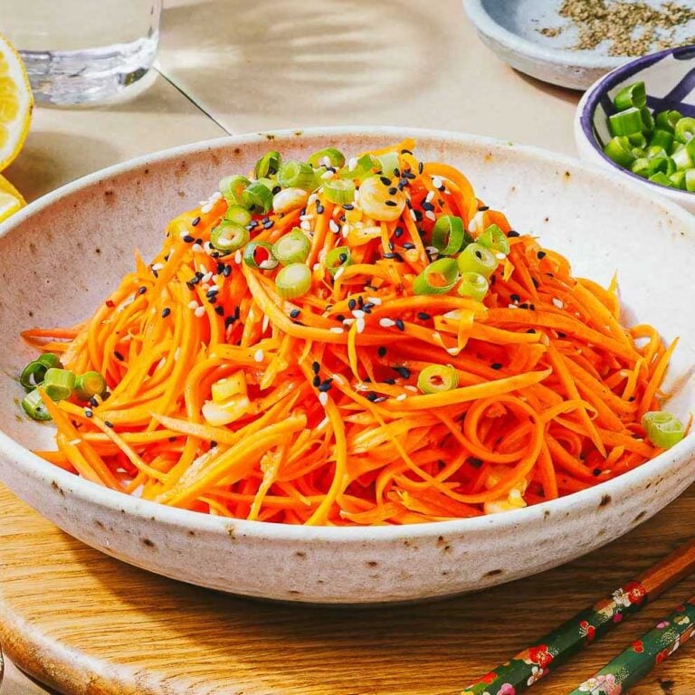 A bowl of Korean carrot salad topped with sliced green onions and black sesame seeds, placed on a wooden surface with chopsticks and small dishes of condiments nearby.