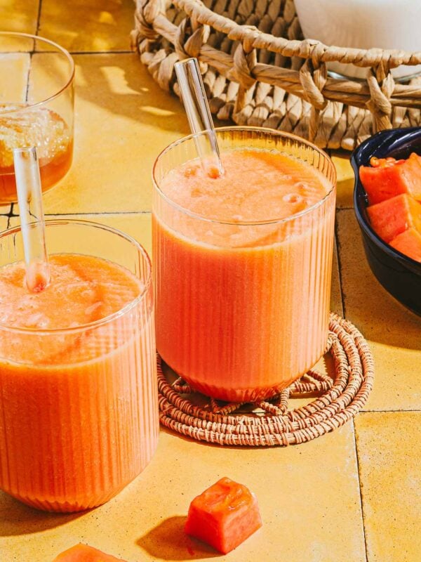 Two glasses of orange papaya milk smoothie with glass straws sit on a yellow tiled surface, surrounded by papaya cubes and a woven basket in the background.