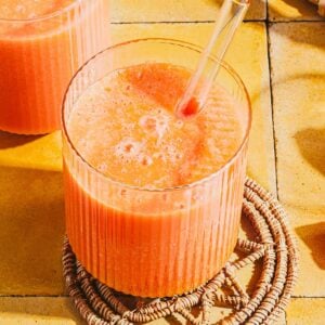 A vibrant papaya milk smoothie in a ribbed glass with a clear straw, placed on a woven coaster atop a yellow tiled surface in bright sunlight.