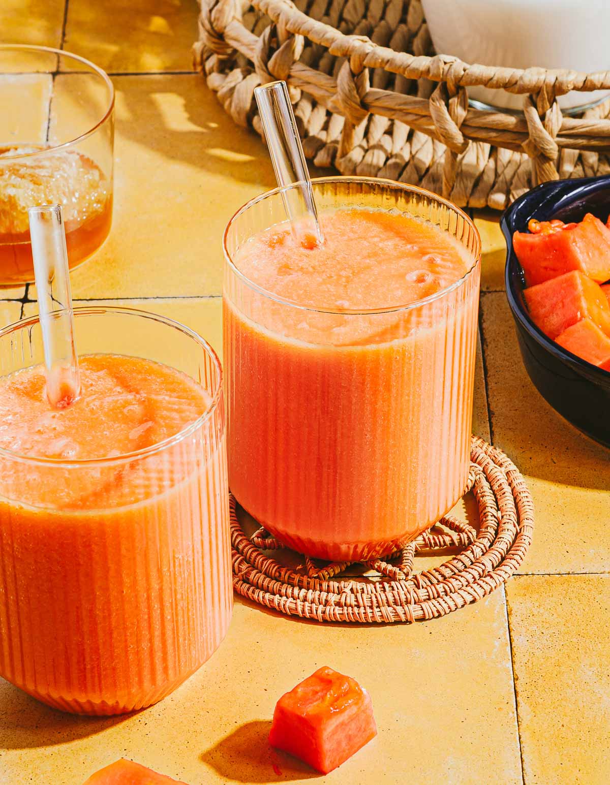 Two glasses of orange papaya milk smoothie with glass straws sit on a yellow tiled surface, surrounded by papaya cubes and a woven basket in the background.