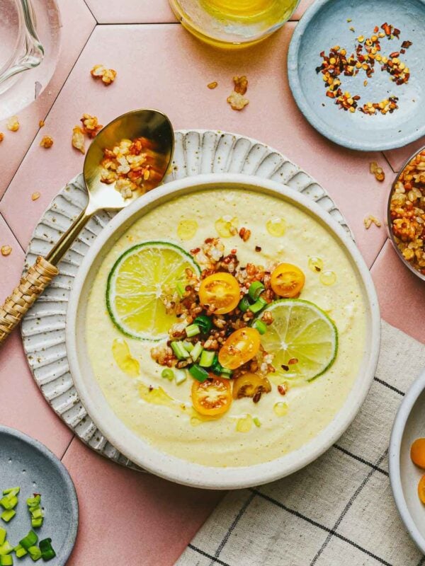 A bowl of creamy yellow Corn Gazpacho topped with lime slices, yellow cherry tomatoes, chopped green onions, and crispy grains, with a spoon and small plates of toppings nearby on a pink tiled surface.