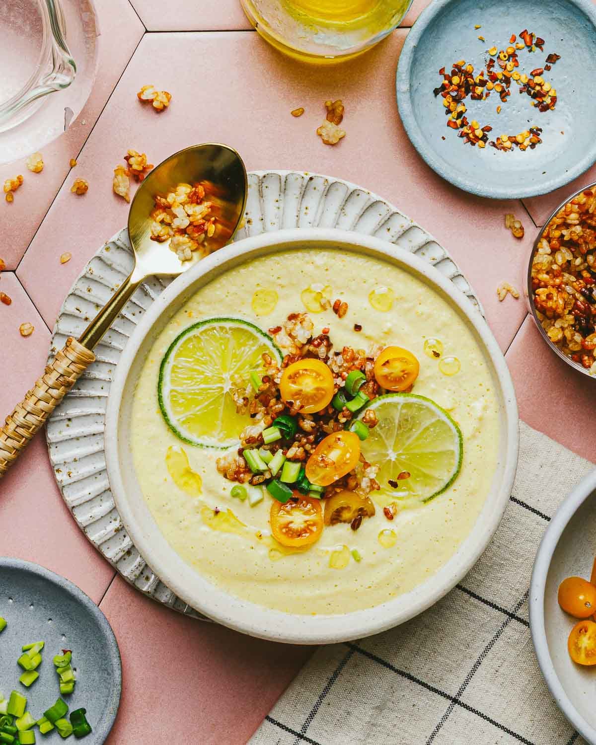 A bowl of creamy yellow Corn Gazpacho topped with lime slices, yellow cherry tomatoes, chopped green onions, and crispy grains, with a spoon and small plates of toppings nearby on a pink tiled surface.
