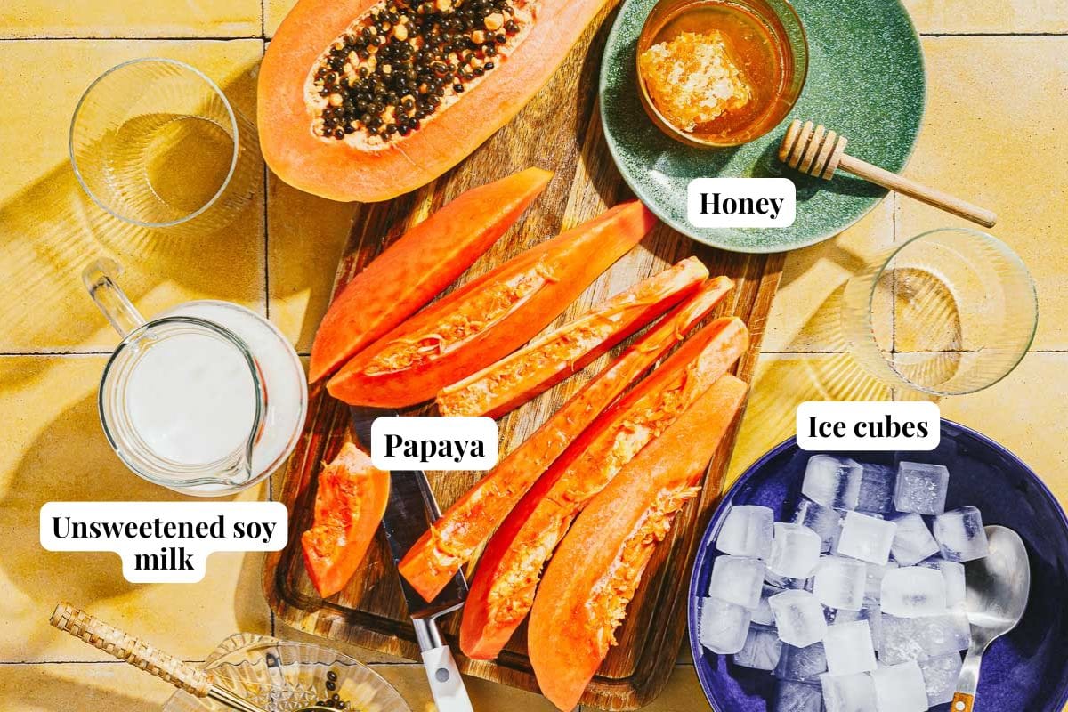 A top-down view of sliced papaya on a wooden board, surrounded by a bowl of ice cubes, a green plate with honey and a honey dipper, and a glass of Papaya Milk. Each item is labeled in the image.