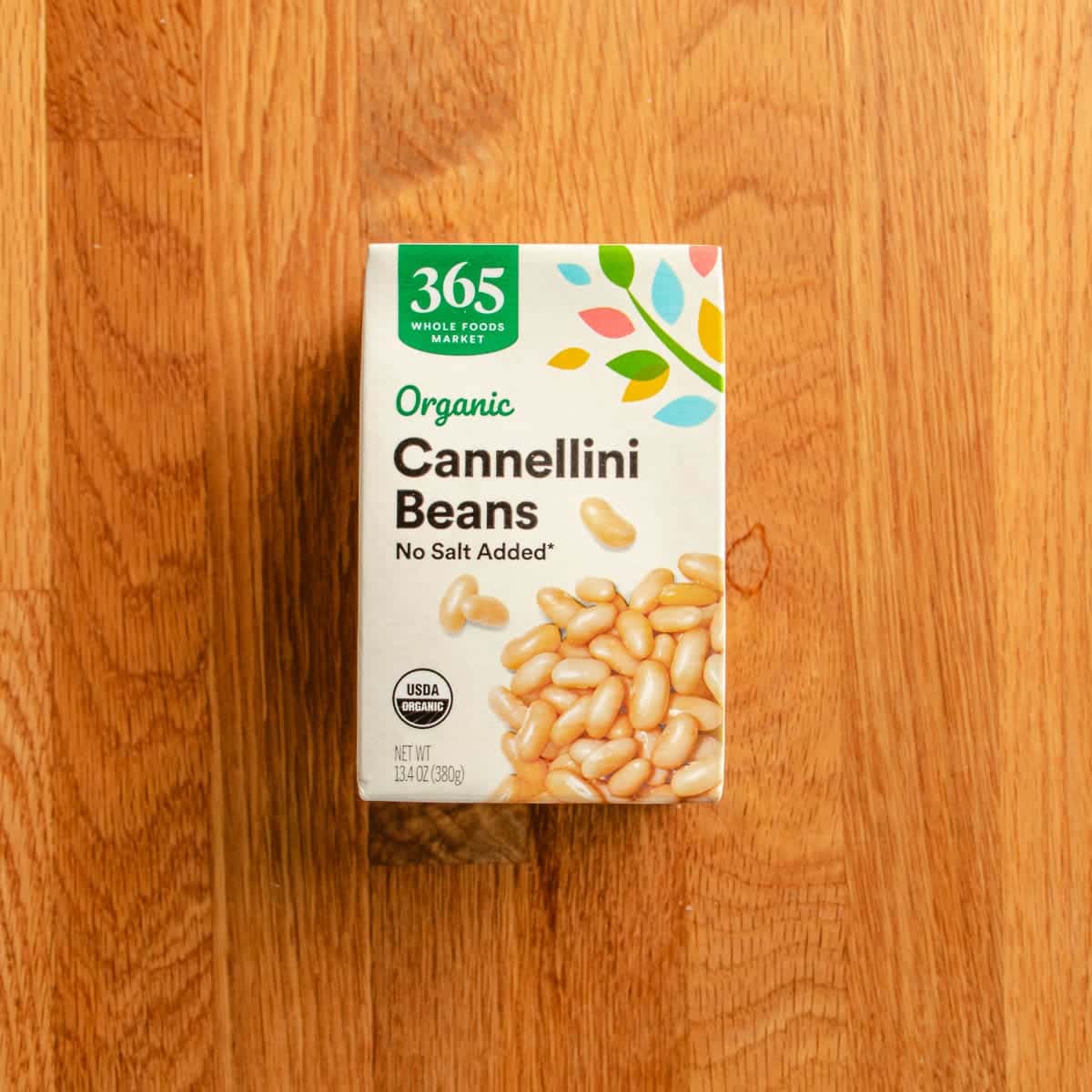 A box of 365 Whole Foods Market Organic Cannellini Beans with No Salt Added sits on a wooden surface. The packaging shows white beans and colorful leaf designs—perfect for adding to your favorite chicken cauliflower casserole.