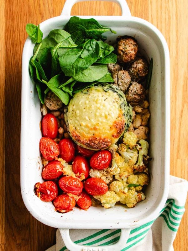 A white baking dish with roasted cherry tomatoes, white beans, chicken meatballs, cauliflower, fresh spinach, and a large stuffed spinach artichoke dip on a wooden surface evokes the comfort of a chicken cauliflower casserole next to a striped cloth.