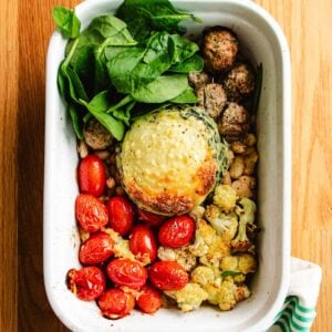 A white dish holds chicken cauliflower casserole, cooked chicken, fresh spinach, roasted cherry tomatoes, white beans, and a baked mound with melted cheese topping—all beautifully arranged on a wooden surface.