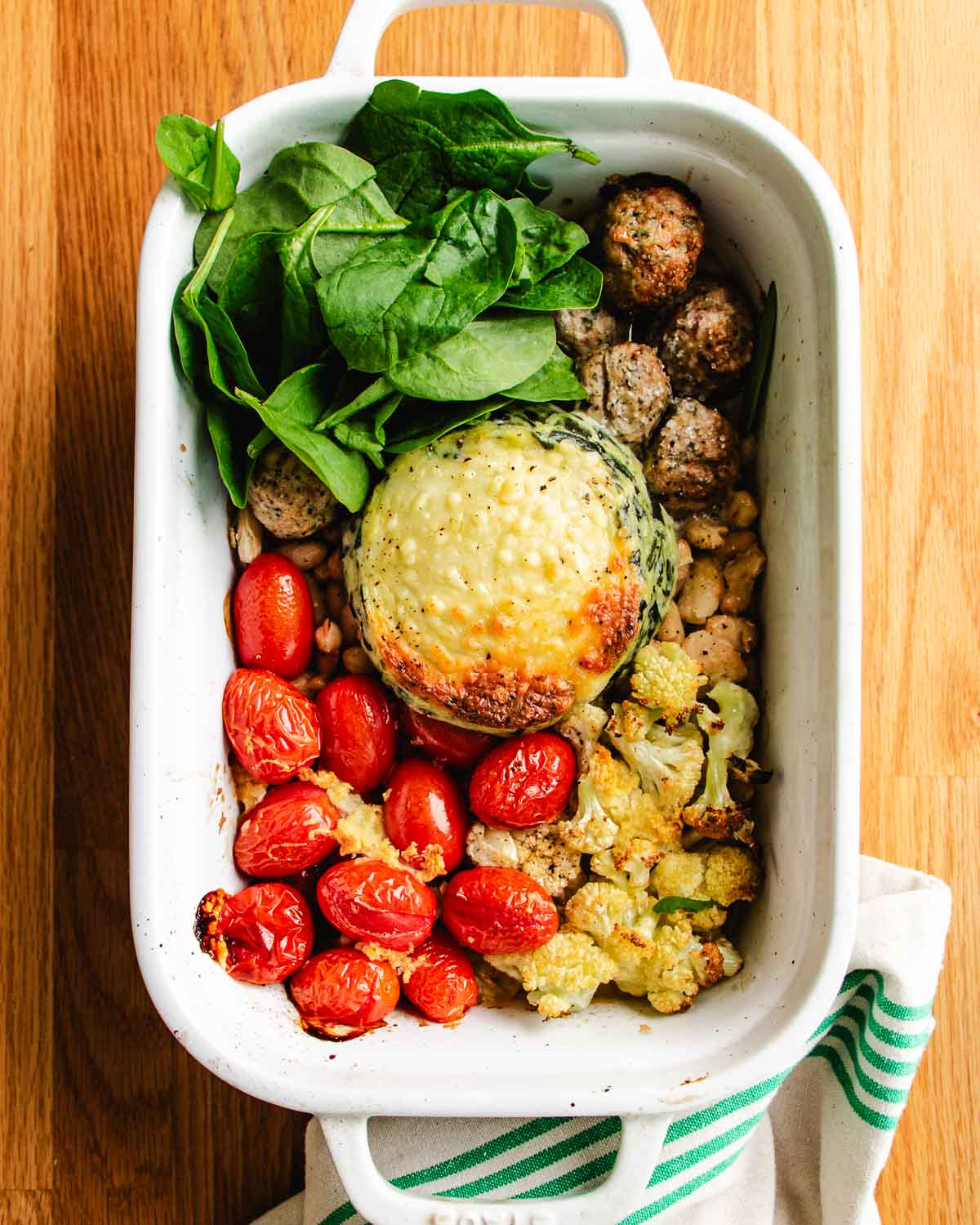 A white baking dish with roasted cherry tomatoes, white beans, chicken meatballs, cauliflower, fresh spinach, and a large stuffed spinach artichoke dip on a wooden surface evokes the comfort of a chicken cauliflower casserole next to a striped cloth.