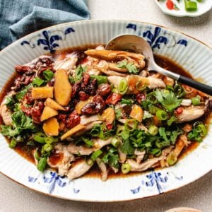 A platter of Chinese steamed chicken topped with sliced ginger, goji berries, fresh herbs, and chopped green onions, served with a savory sauce. A spoon rests on the plate, and garnishes are nearby.