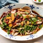 A platter of Chinese steamed chicken topped with sliced ginger, goji berries, dates, chopped green onions, and fresh herbs in a soy-based sauce, served with a spoon.
