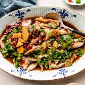 A platter of Chinese steamed chicken topped with sliced ginger, goji berries, dates, chopped green onions, and fresh herbs in a soy-based sauce, served with a spoon.
