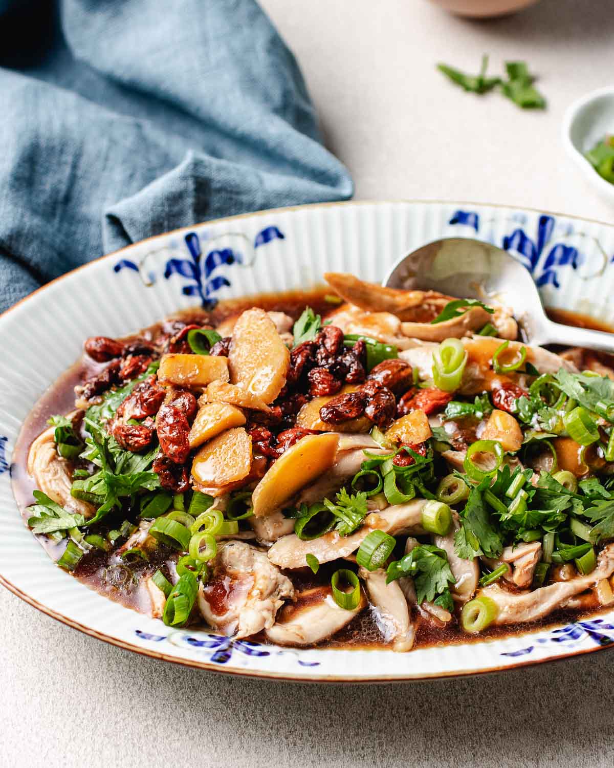 A plate of Chinese steamed chicken topped with goji berries and dates, chopped green onions, and herbs, served in a savory sauce with a spoon on the side and a blue napkin in the background.