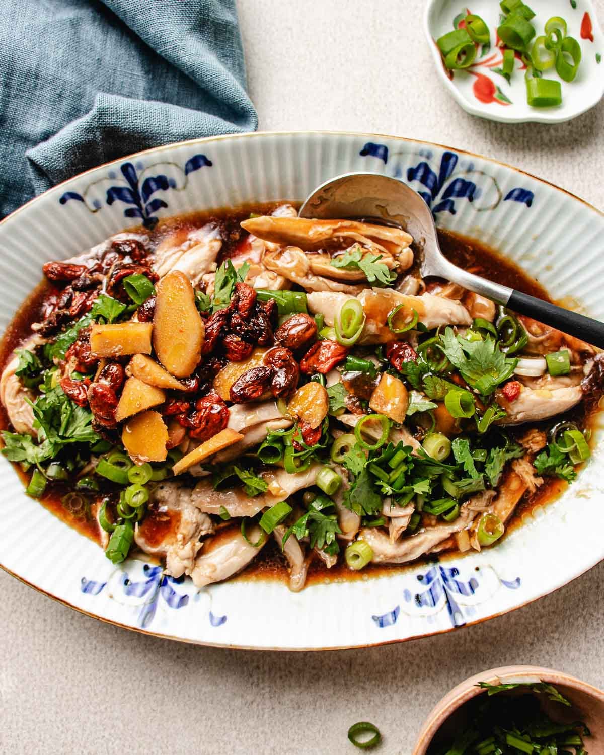 A platter of Chinese steamed chicken topped with sliced ginger, goji berries, fresh herbs, and chopped green onions, served with a savory sauce. A spoon rests on the plate, and garnishes are nearby.