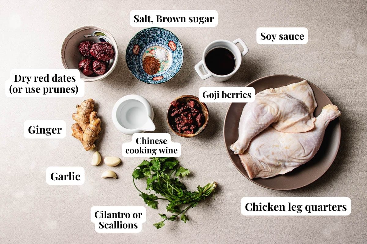 Top-down view of labeled ingredients for Chinese steamed chicken: chicken leg quarters, soy sauce, goji berries, dry red dates or prunes, ginger, garlic, cilantro or scallions, Chinese cooking wine, salt, and brown sugar.