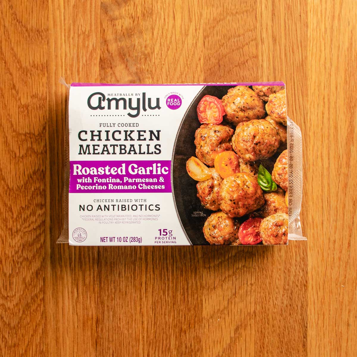 A package of Amylu fully cooked chicken meatballs in roasted garlic flavor sits on a wooden surface—perfect for adding to your favorite chicken cauliflower casserole. The label highlights fontina, parmesan, pecorino romano cheeses, and no antibiotics.