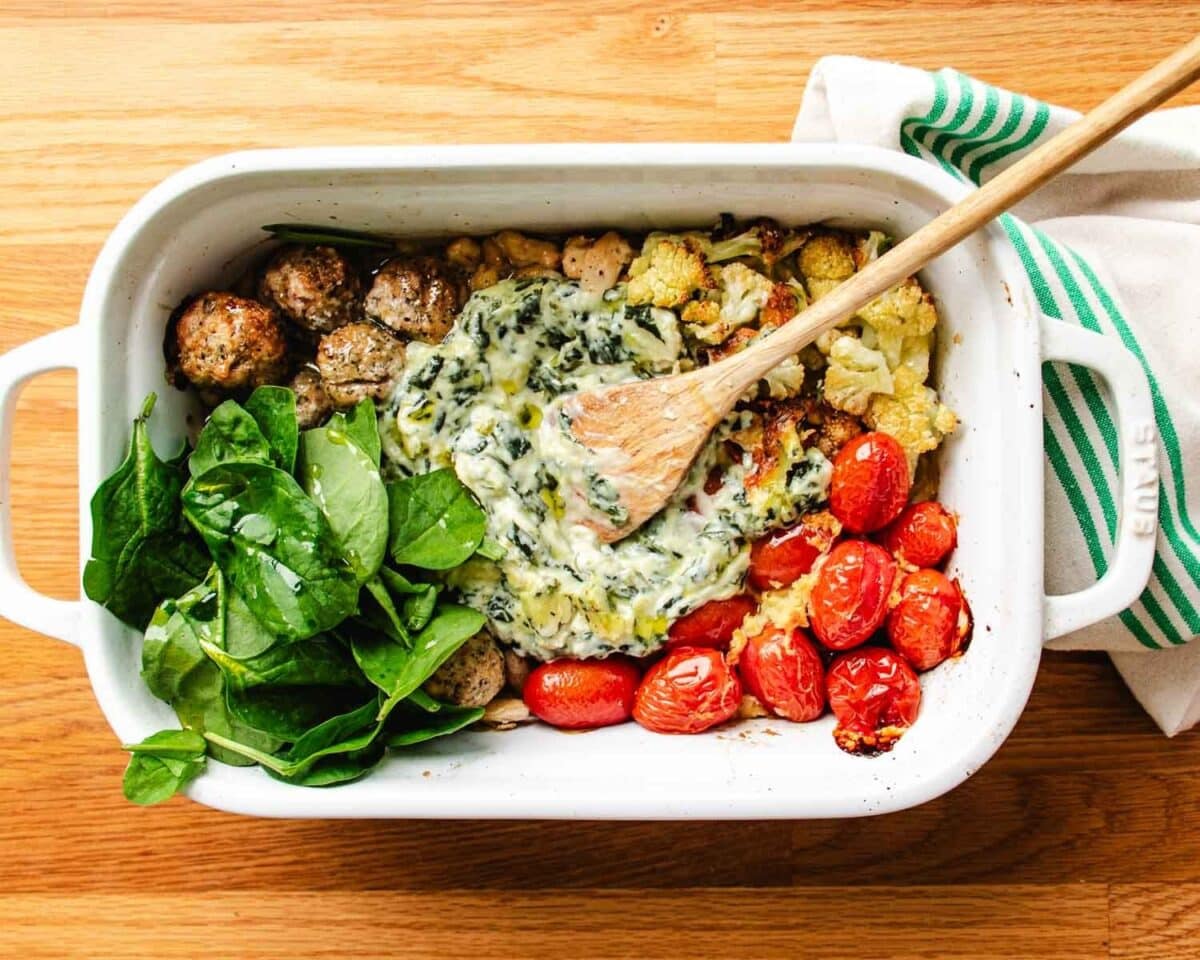 A white baking dish with spinach leaves, roasted meatballs, blistered cherry tomatoes, cooked chicken, cauliflower, and creamy spinach artichoke dip—like a deconstructed chicken cauliflower casserole—with a wooden spoon and a striped kitchen towel on the side.