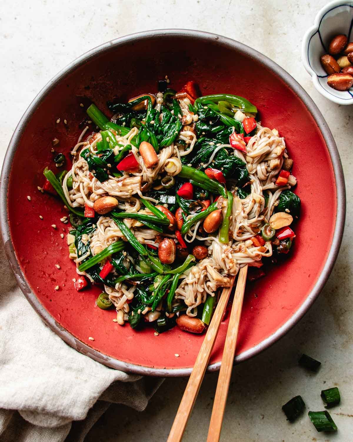 A vibrant cold enoki mushroom salad with spinach, red peppers, and roasted peanuts, tossed in a garlicky, tangy soy-vinegar dressing and served in a deep red bowl.
