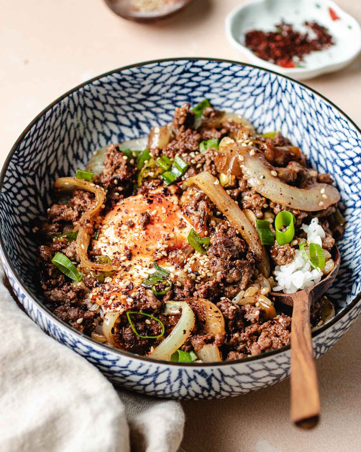 A patterned blue-and-white bowl filled with ground beef bulgogi, sautéed onions, a soft-cooked egg, white rice, chopped green onions, sesame seeds, and red pepper flakes, with a wooden spoon resting inside.