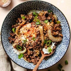 A blue and white bowl loaded with ground beef bulgogi, soft simmered bulgogi sauce eggs, sweet onions, green onions over a bed of steamed white rice with a wooden spoon on the side for serving.