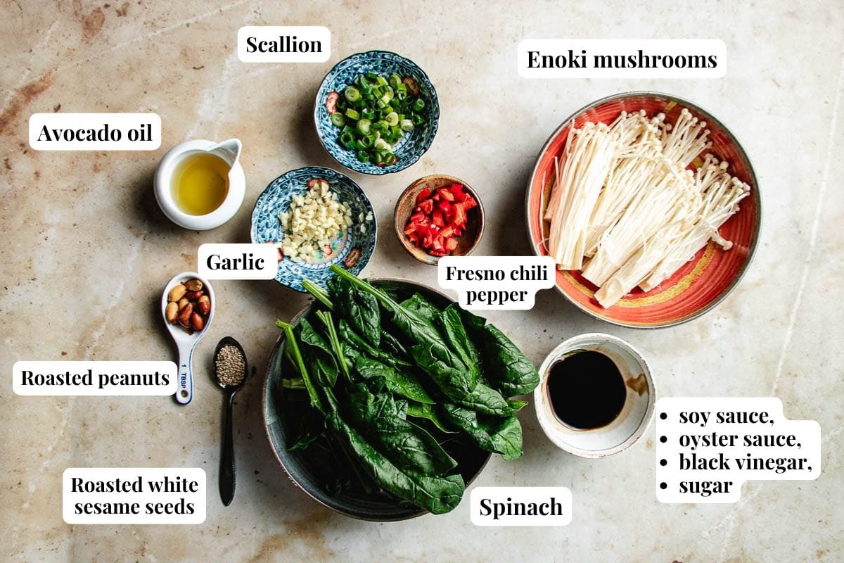 Various labeled ingredients for an Enoki mushroom salad are arranged on a surface, including enoki mushrooms, scallions, spinach, garlic, Fresno chili pepper, avocado oil, roasted peanuts, sesame seeds, soy sauce, and condiments.