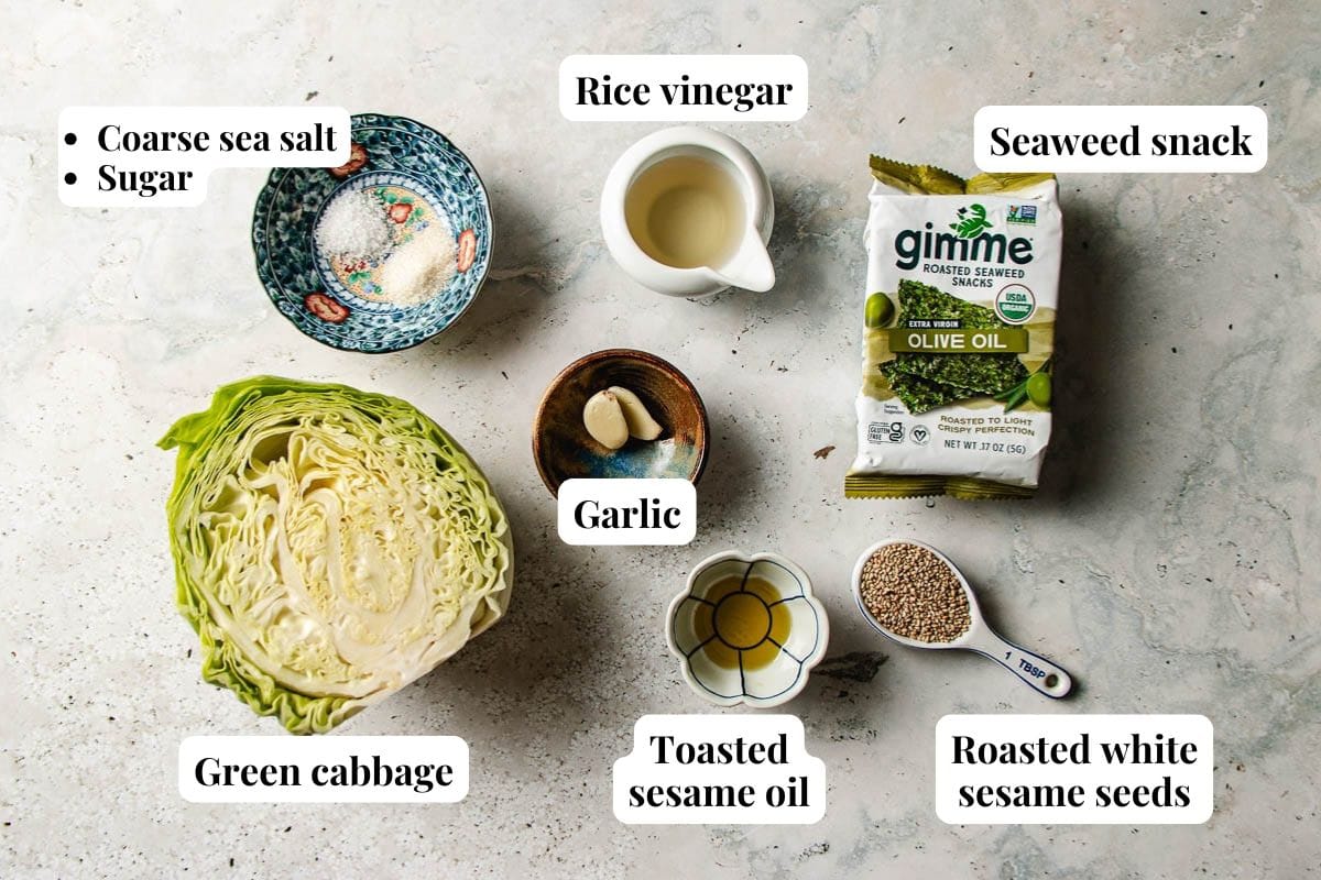 Top-down view of labeled ingredients for Korean cabbage salad: coarse sea salt, sugar, rice vinegar, seaweed snack, green cabbage, toasted sesame oil, garlic cloves, and roasted white sesame seeds on a light surface.