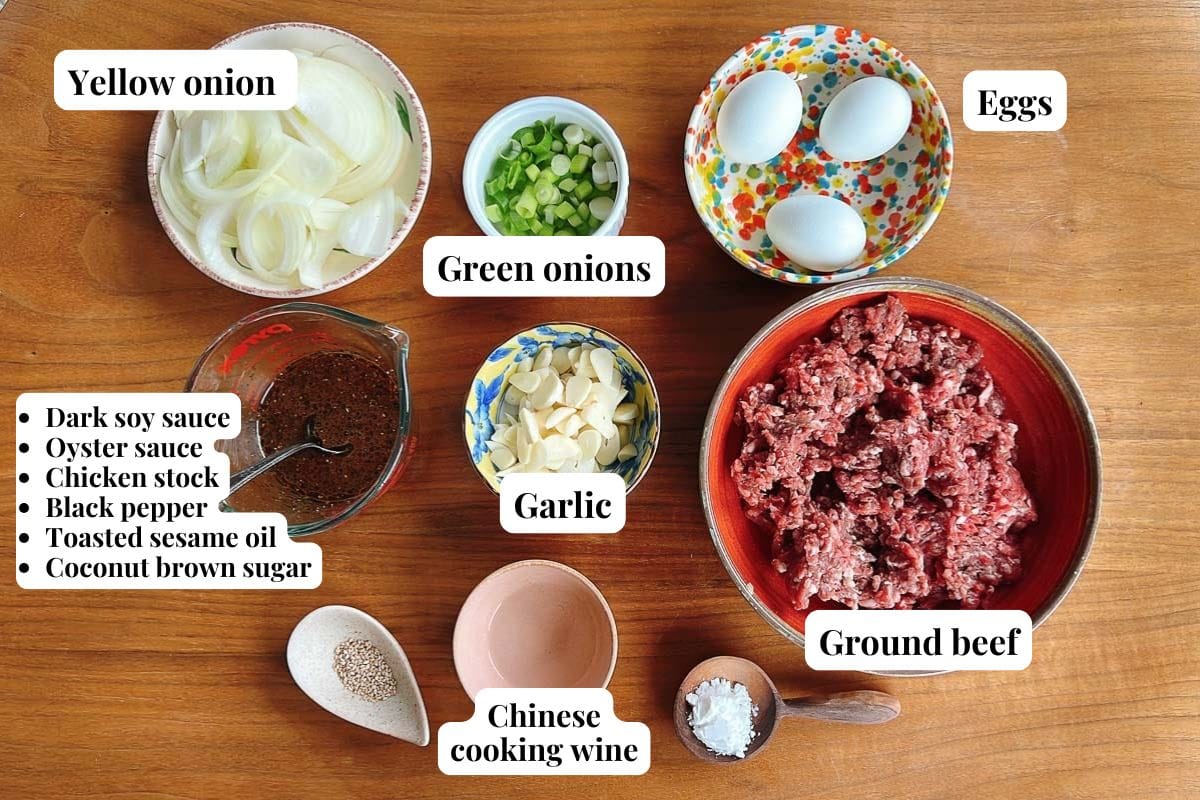 A top-down view of labeled ingredients for ground beef bulgogi in bowls and cups: sliced yellow onion, green onions, eggs, minced garlic, ground beef, Chinese cooking wine, soy sauce, oyster sauce, chicken stock, black pepper, sesame oil, and brown sugar.