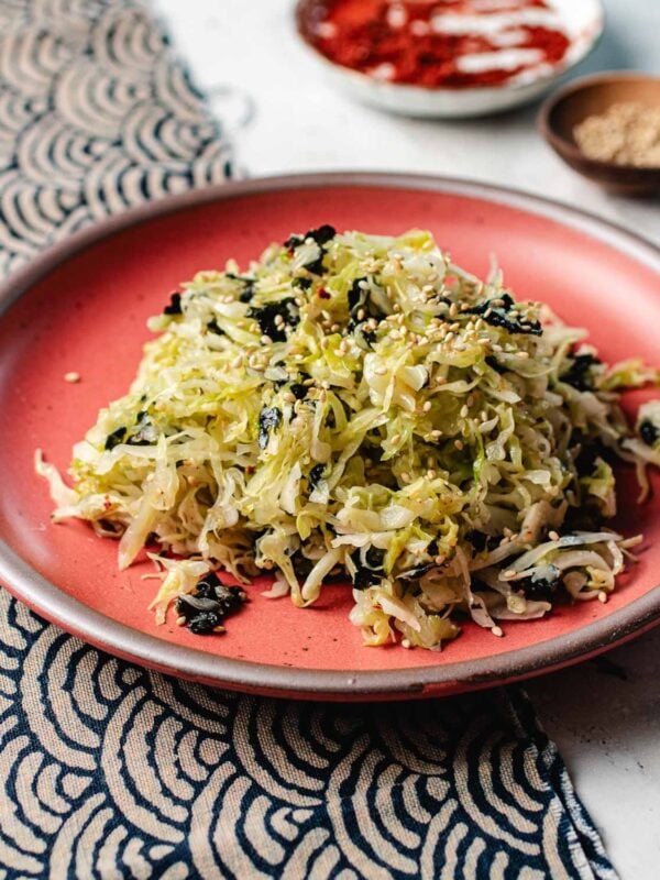 A red plate holds a mound of Korean cabbage salad mixed with seaweed and sprinkled with sesame seeds. In the background, small bowls with spices and seeds rest on a patterned cloth.