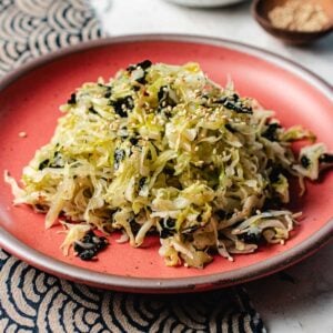 A mound of Korean cabbage salad mixed with seaweed and sprinkled with sesame seeds sits on a red plate, set atop a table with a patterned cloth.