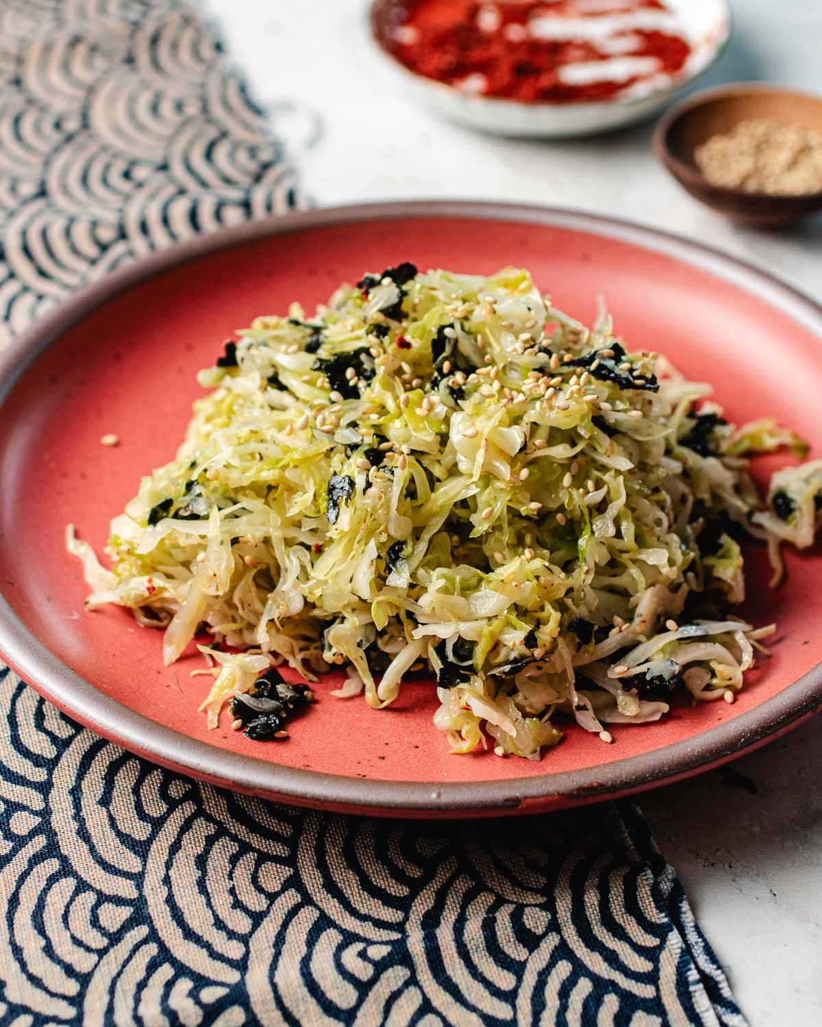 A red plate holds a mound of Korean cabbage salad mixed with seaweed and sprinkled with sesame seeds. In the background, small bowls with spices and seeds rest on a patterned cloth.