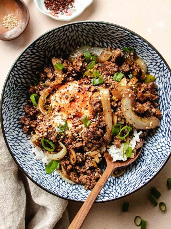 A patterned bowl filled with white rice, ground beef bulgogi, sautéed onions, a bulgogi sauce simmered egg, sliced green onions, and a sprinkle of sesame seeds, with a wooden spoon resting inside.