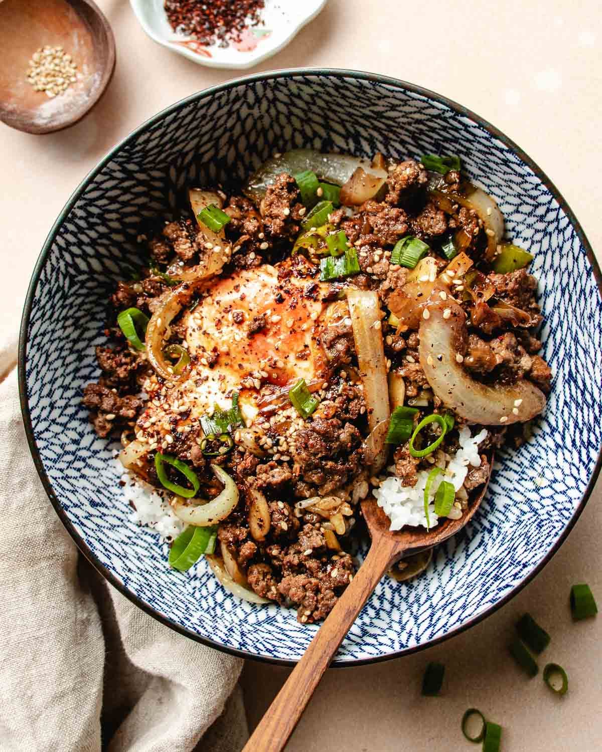A patterned bowl filled with white rice, ground beef bulgogi, sautéed onions, a bulgogi sauce simmered egg, sliced green onions, and a sprinkle of sesame seeds, with a wooden spoon resting inside.