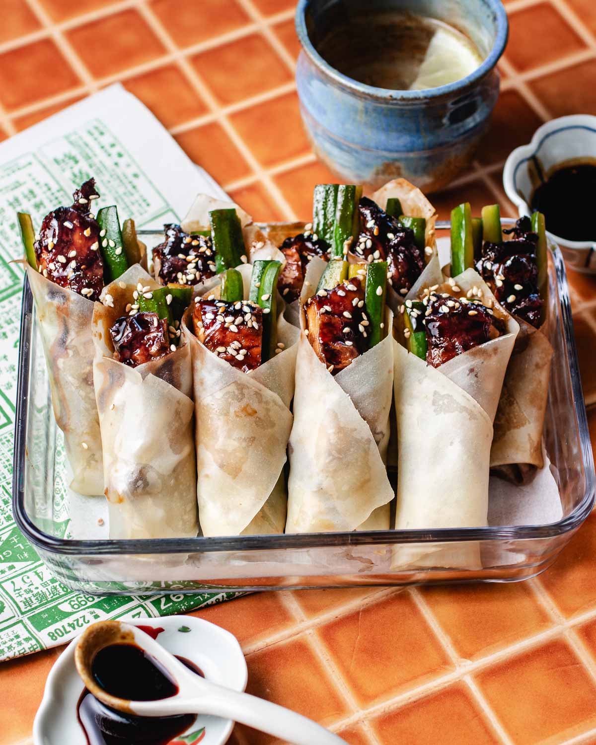 A glass dish filled with neatly wrapped soy sauce chicken spring rolls, garnished with sesame seeds and sliced cucumbers, sits on a tiled table beside bowls of dipping sauce—including a soy sauce chicken marinade—and a ceramic cup.