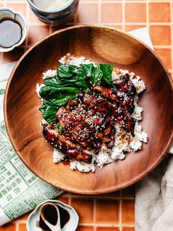 A wooden bowl filled with white rice, glazed caramelized meat slices made with a soy sauce chicken marinade, topped with sesame seeds, chopped green bok choy, and red chili. Small dishes of soy sauce and a napkin are nearby on a tiled surface.