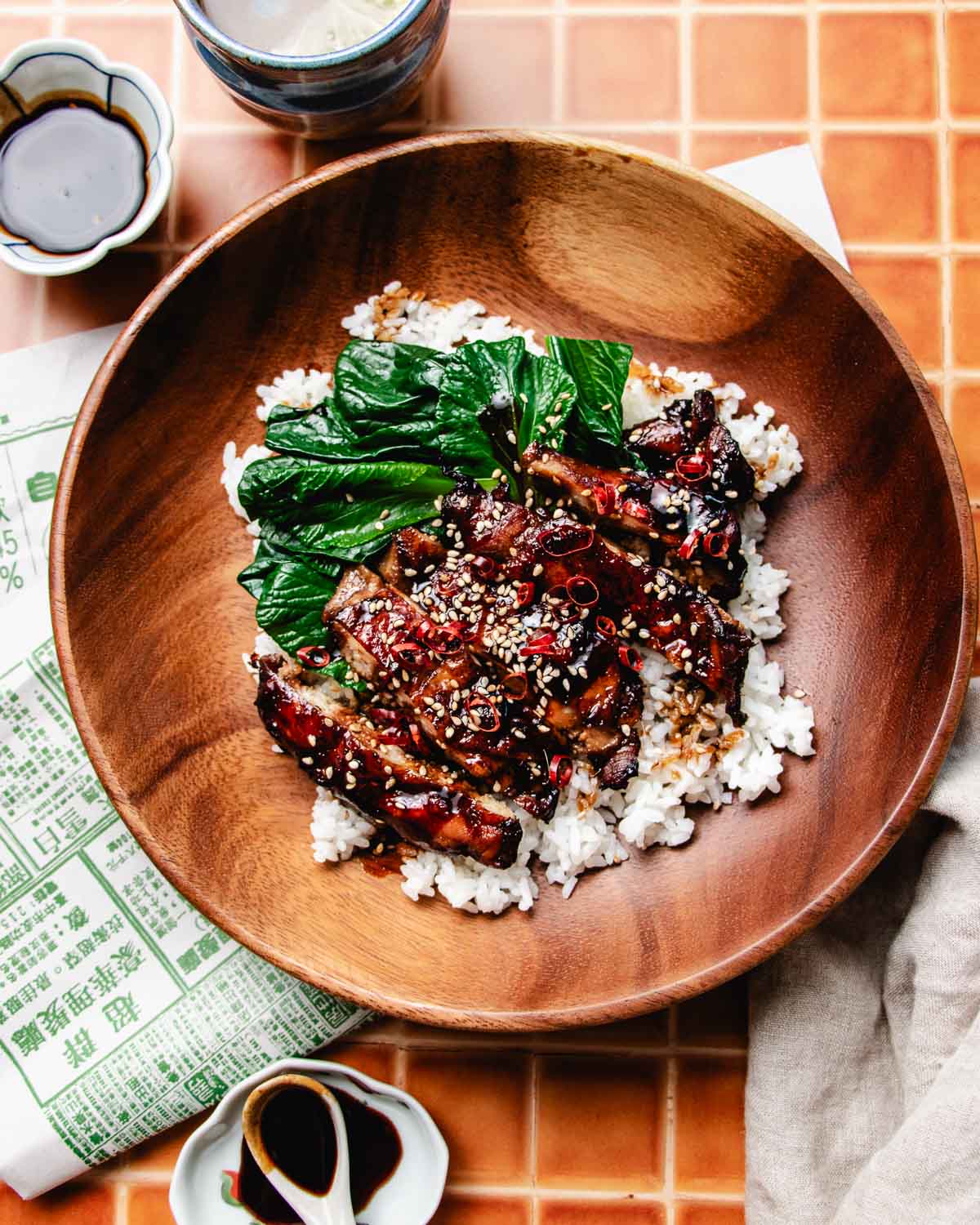 A wooden bowl filled with white rice, glazed caramelized meat slices made with a soy sauce chicken marinade, topped with sesame seeds, chopped green bok choy, and red chili. Small dishes of soy sauce and a napkin are nearby on a tiled surface.