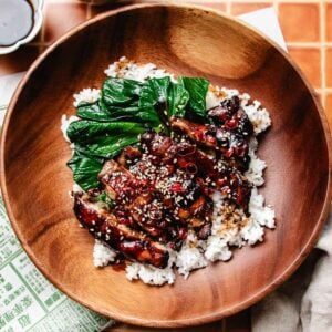 A wooden bowl filled with white rice, topped with glazed caramelized chicken meat slices marinated in soy sauce chicken marinade, leafy greens, and garnished with sesame seeds and sliced red chili peppers.
