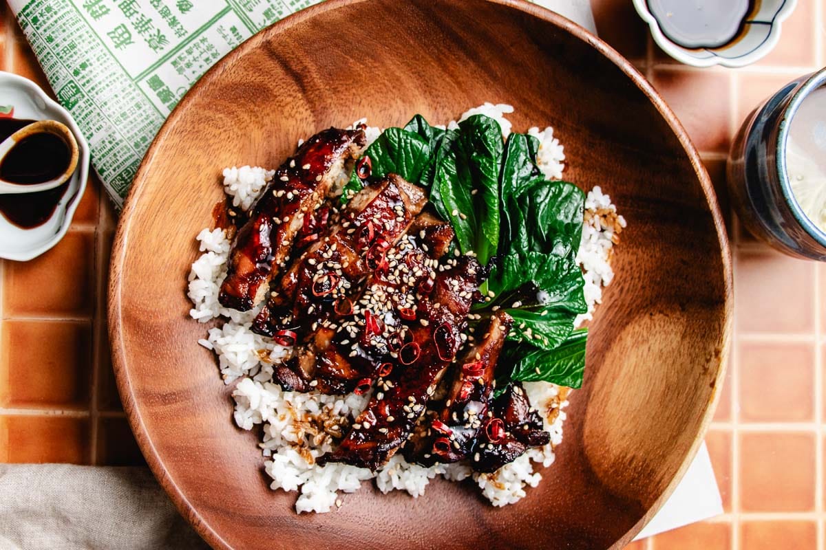 A wooden bowl filled with white rice, topped with glazed soy chicken meat marinated in soy sauce chicken marinade, leafy greens, sliced red chili, and sesame seeds, placed on a tiled surface with sauces and a drink nearby.