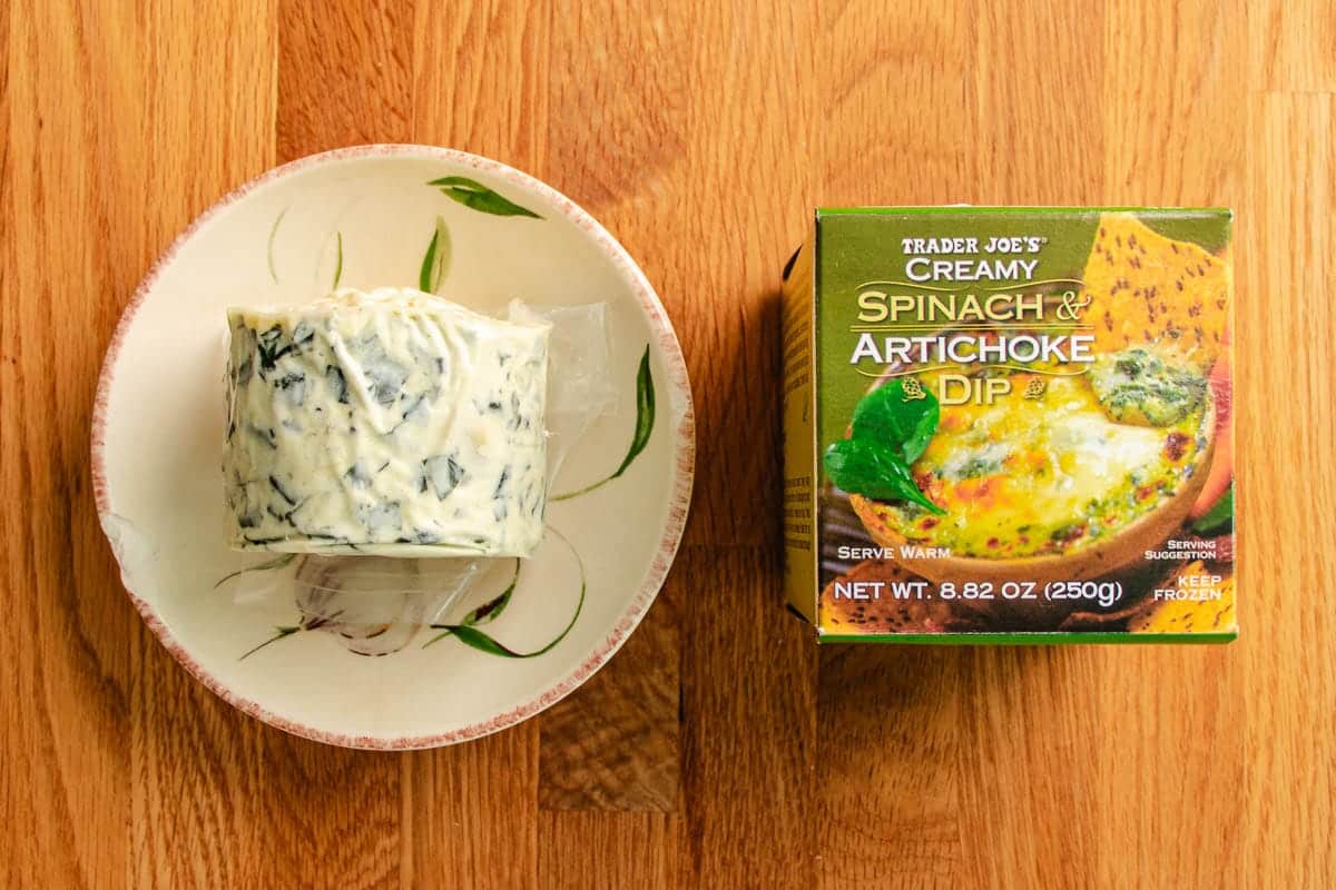 A round block of packaged spinach and artichoke dip sits on a plate next to a Trader Joe’s creamy spinach & artichoke dip box, easy quick way to flavor the hearty chicken cauliflower casserole.