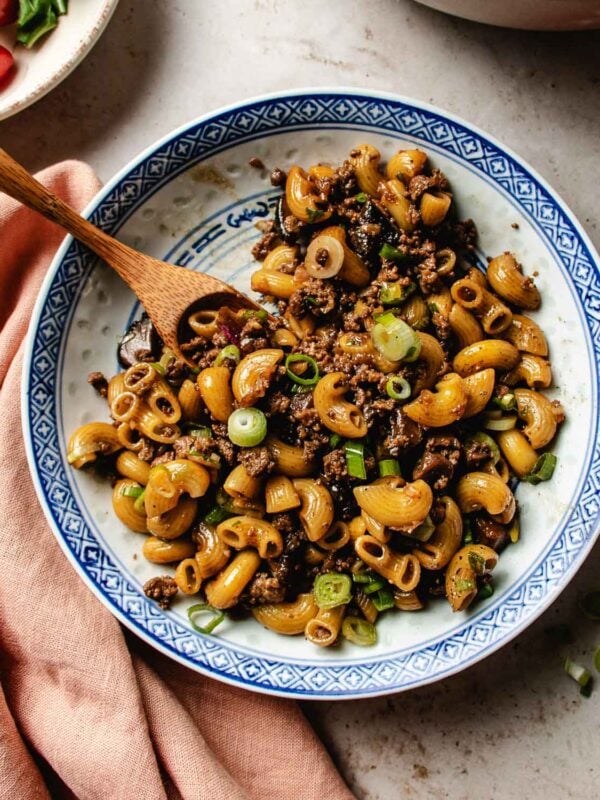 A patterned blue and white bowl filled with ground beef macaroni, garnished with chopped green onions, with a wooden spoon resting inside the bowl.