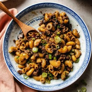 A blue-and-white bowl filled with ground beef macaroni, chopped green onions, and macaroni noodles, with a wooden spoon resting inside. The bowl sits on a light surface with a peach-colored cloth nearby.