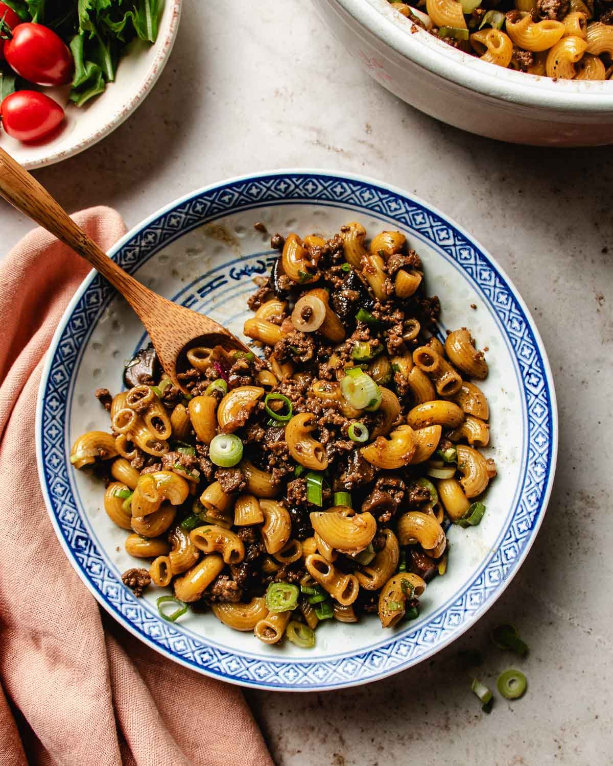 A bowl of ground beef macaroni pasta mixed with ground meat and topped with chopped green onions, served with a wooden spoon on a light-colored surface next to a pink napkin.