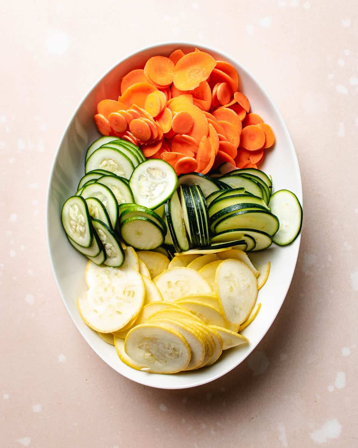 An oval white dish filled with neatly arranged slices of carrots, green zucchini, and yellow squash on a light, speckled background resembles the colorful presentation often found in Bulgogi Bibimbap.