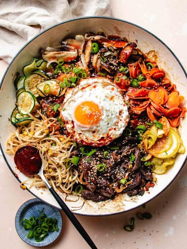 A large bowl of Bulgogi Bibimbap with sliced beef, vegetables, noodles, a sunny-side-up egg, and a dollop of red sauce, garnished with sesame seeds and green onions. A fork and a small dish of chopped green onions are beside it.