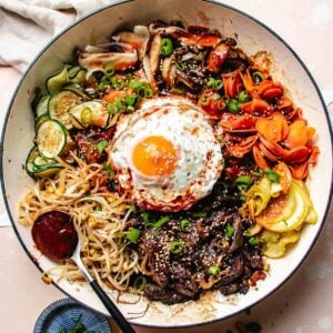 A colorful bowl of Bulgogi Bibimbap features rice, sliced beef, sautéed vegetables, mushrooms, carrots, zucchini, bean sprouts, gochujang sauce, and a fried egg topped with scallions and sesame seeds.