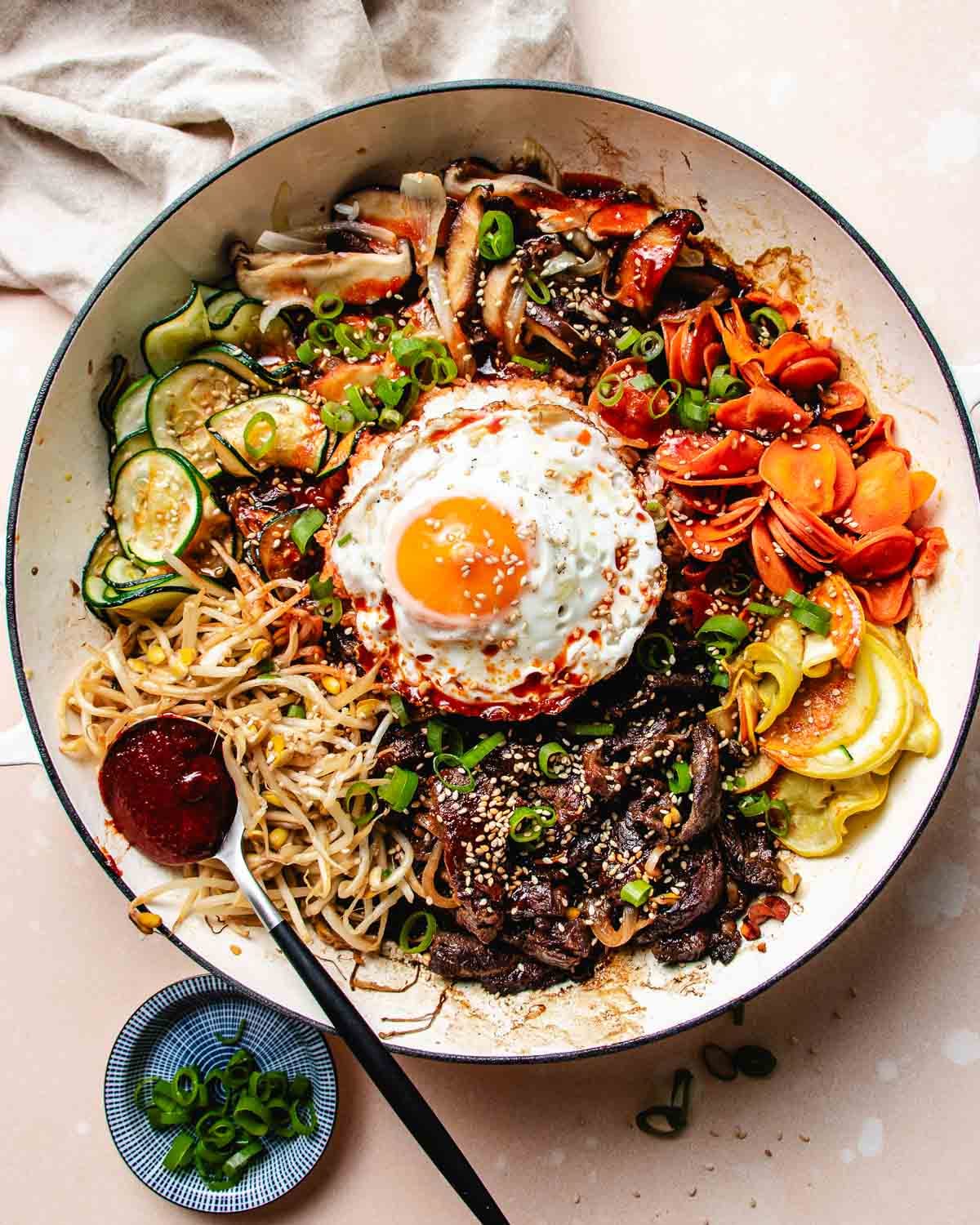 A large bowl of Bulgogi Bibimbap with sliced beef, vegetables, noodles, a sunny-side-up egg, and a dollop of red sauce, garnished with sesame seeds and green onions. A fork and a small dish of chopped green onions are beside it.