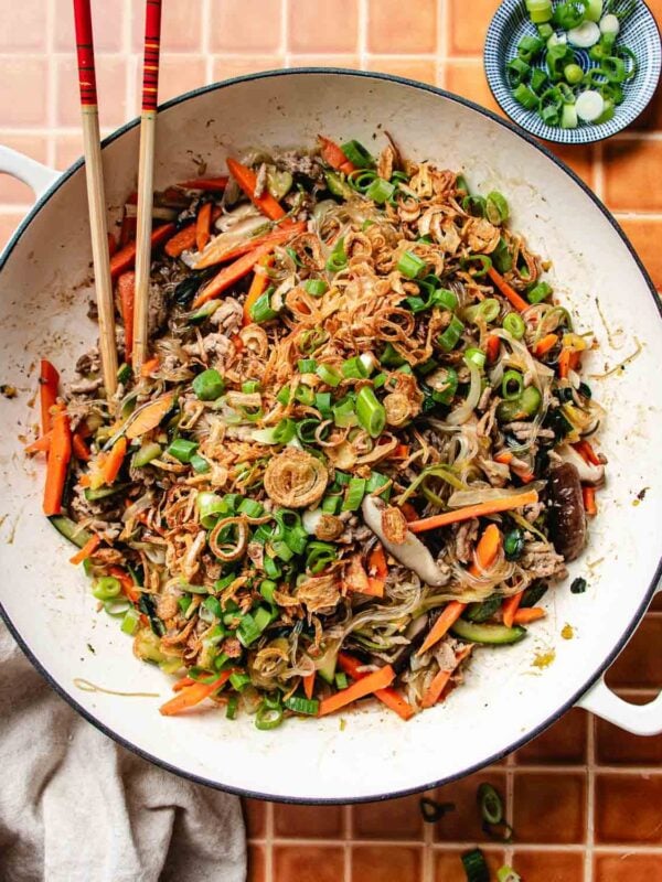A large white pan filled with one pan glass noodles, stir-fried with sliced carrots, mushrooms, green onions, and fried shallots. Chopsticks rest on the rim, with a small bowl of chopped green onions beside the pan.