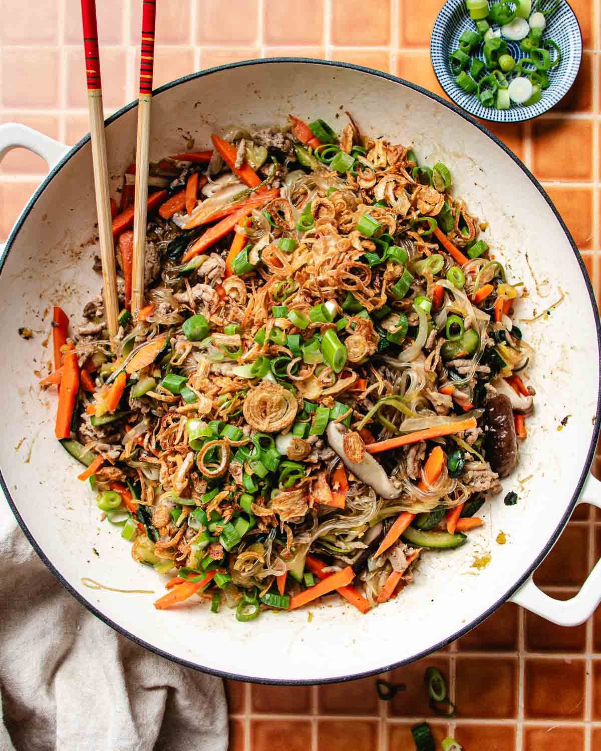 A large white pan filled with one pan glass noodles, stir-fried with sliced carrots, mushrooms, green onions, and fried shallots. Chopsticks rest on the rim, with a small bowl of chopped green onions beside the pan.