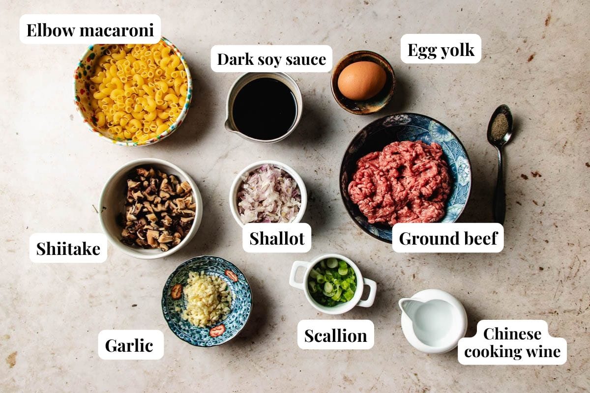 A top-down view of labeled ingredients in bowls for ground beef macaroni: elbow macaroni, dark soy sauce, egg yolk, ground beef, shallot, shiitake mushrooms, garlic, scallion, and Chinese cooking wine on a light surface.