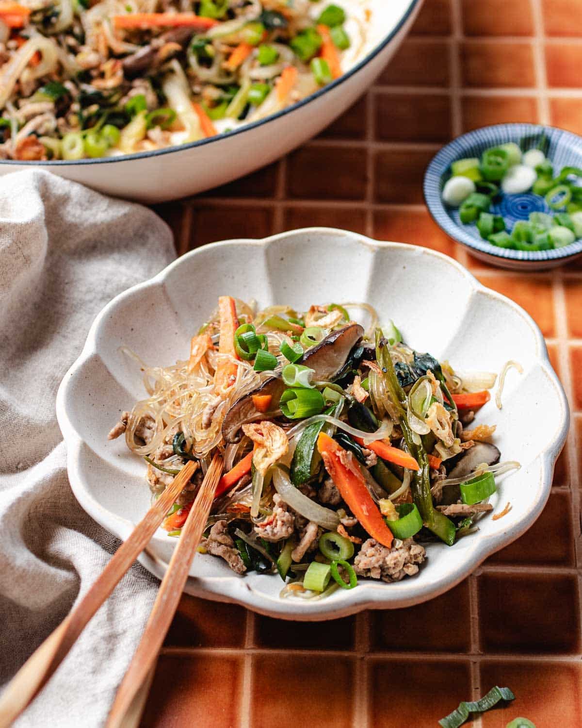 A bowl of mung bean noodles with vegetables and beef, made in one pan with glass noodles, garnished with chopped green onions and served with chopsticks. In the background are a larger dish of japchae and a small bowl of green onions.
