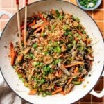 A large white pan filled with one pan glass noodles, stir-fried with sliced carrots, mushrooms, and green onions, then topped with crispy shallots. Chopsticks rest on the side of the pan and a small bowl of sliced green onions is nearby.