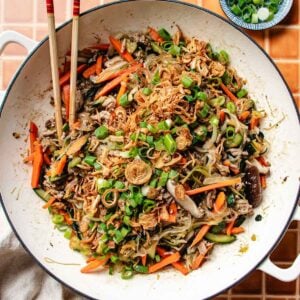 A large white pan filled with one pan glass noodles, stir-fried with sliced carrots, mushrooms, and green onions, then topped with crispy shallots. Chopsticks rest on the side of the pan and a small bowl of sliced green onions is nearby.