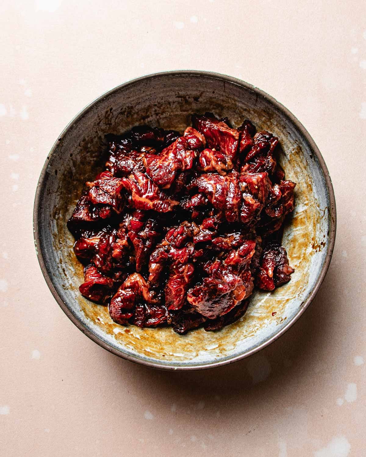 A ceramic bowl filled with marinated beef slices, reminiscent of Bulgogi Bibimbap, is coated in a dark red sauce and sits on a light pink surface with subtle white spots.